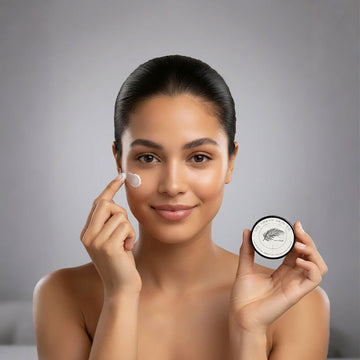 Woman applying cream to her face with a jar of cream in her other hand against a gray background